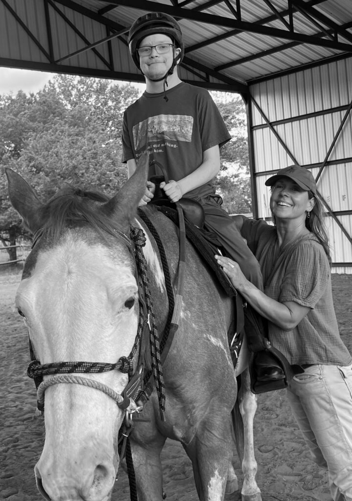 Black and white image of equine therapist Nicol Ragland helping a young client on horseback.
